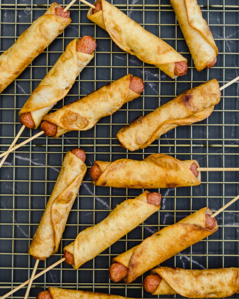 A metal cooling grid on which tortilla-coated hot dogs sausages are placed. The hot dogs are held in place by wooden skewers.