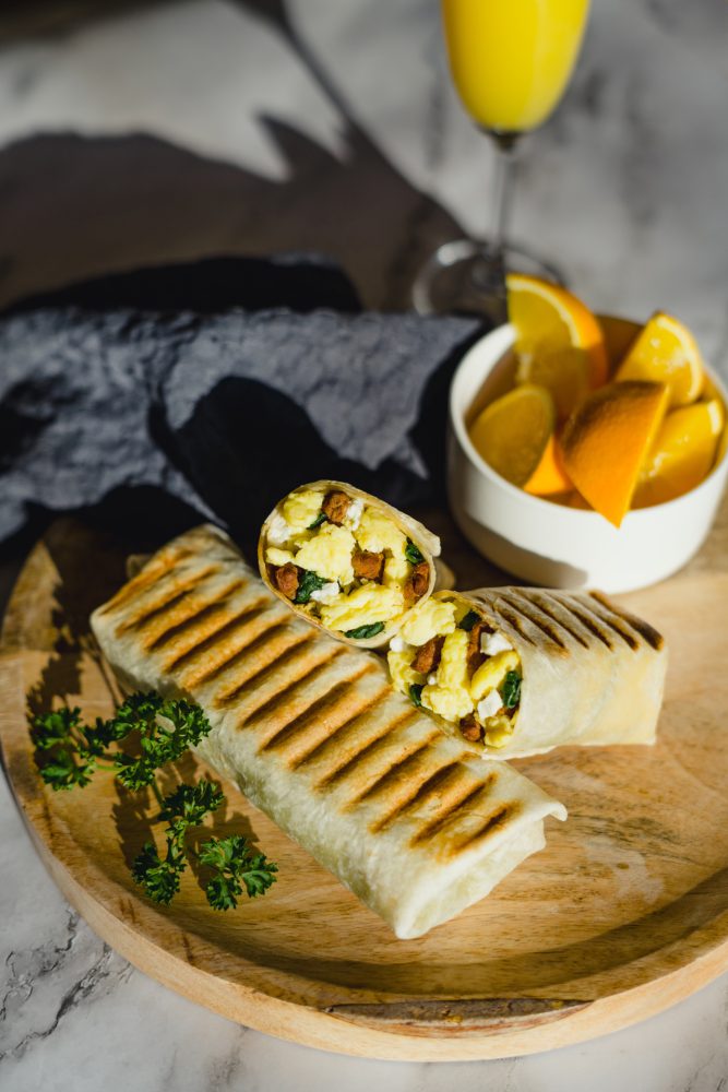 A breakfast burrito cut in half on a wooden board, revealing a filling of scrambled eggs, sausage, spinach and feta cheese. A glass of orange juice and orange wedges are arranged alongside.