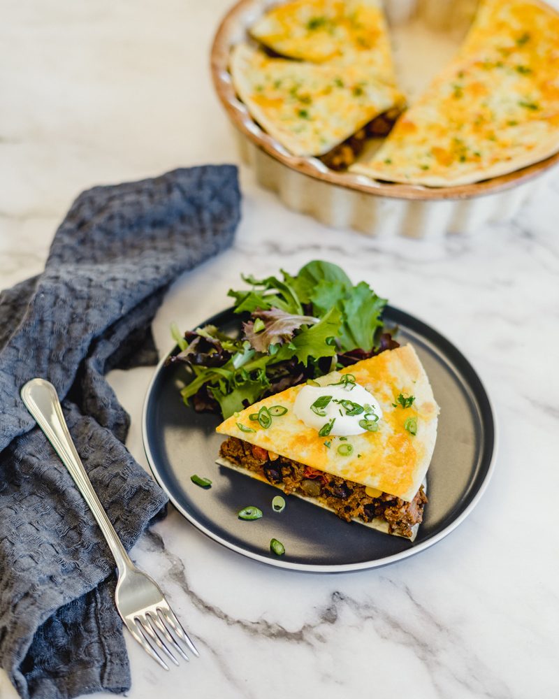 A slice of Mexican pie on a gray plate with a green salad. The pie is made with tortillas, ground beef, vegetables and cheese. The rest of the pie is in a pie plate in the background.