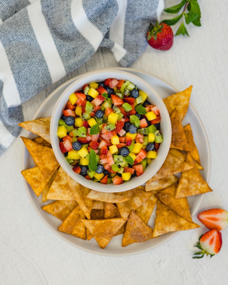 A white bowl filled with fresh fruit salsa, including strawberries, mangoes, kiwis and blueberries, sits on a plate filled with baked tortilla chips sprinkled with cinnamon sugar. Whole strawberries and a blue-and-white-striped napkin are arranged next to the plate.