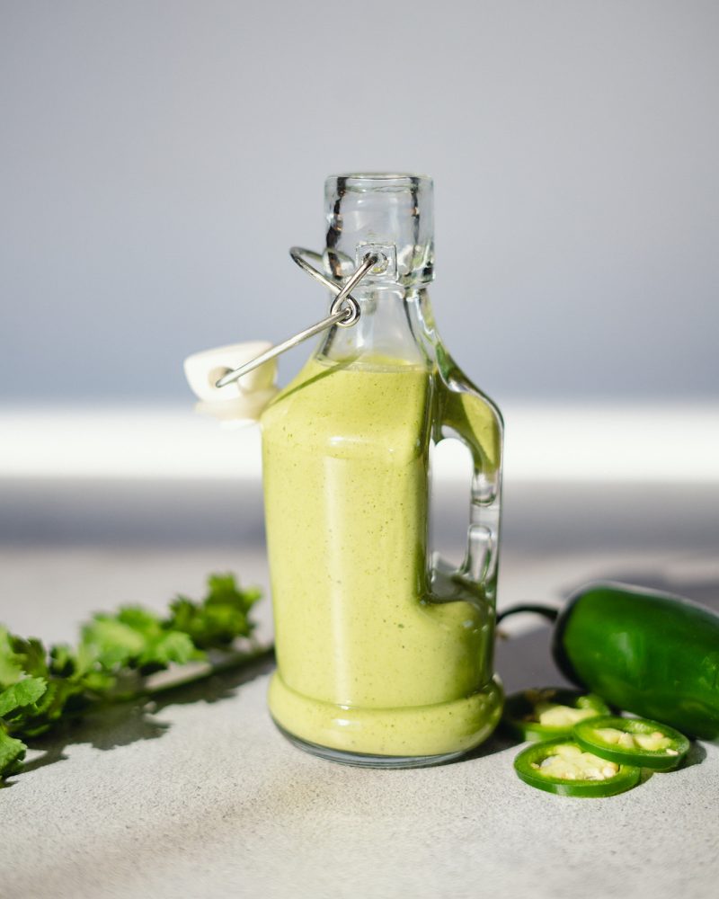 A glass bottle filled with a creamy green sauce, probably a creamy Mexican avocado sauce. The bottle is surrounded by fresh cilantro and slices of jalapeño, key ingredients in the sauce.