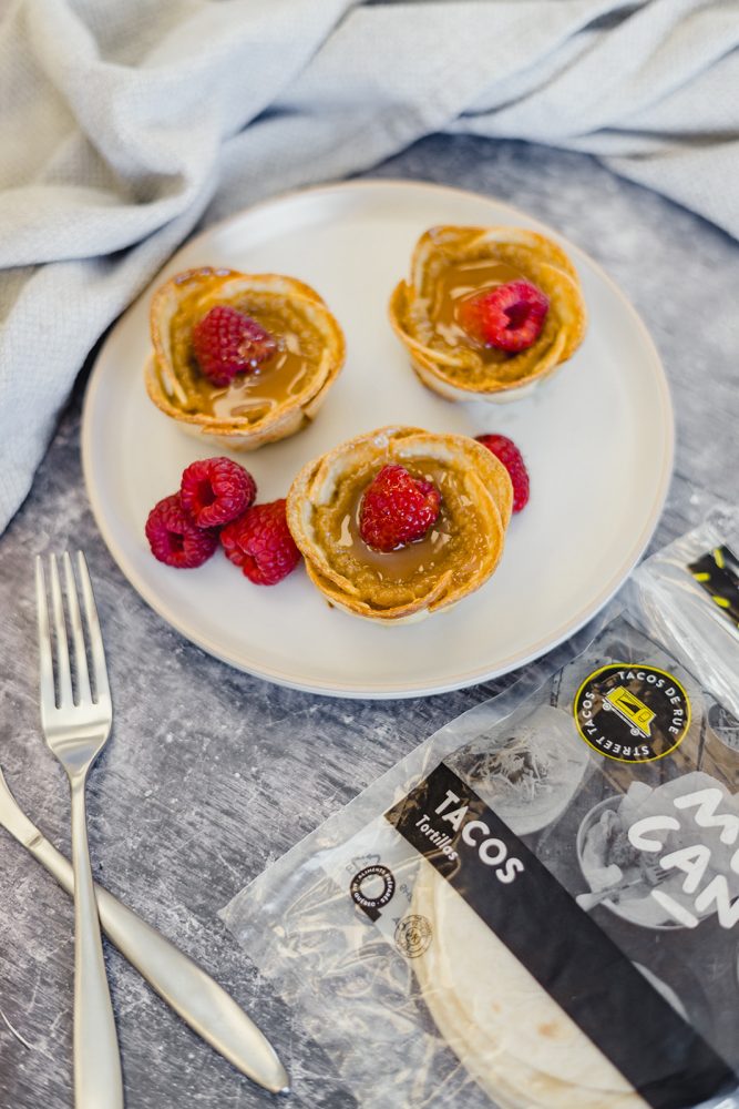 Three mini cream sugar tarts on a white plate, topped with fresh raspberries. A pack of "Street Tacos" tortillas sits next to them, suggesting that the tartlets were made with tortillas. Two forks are laid out on the table.