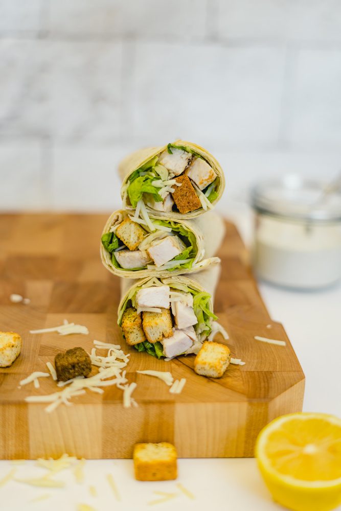 Three chicken Caesar wraps stacked on a wooden cutting board, topped with romaine lettuce, grilled chicken, parmesan and croutons. Additional ingredients are scattered around the wraps, including grated Parmesan, croutons and half a lemon.