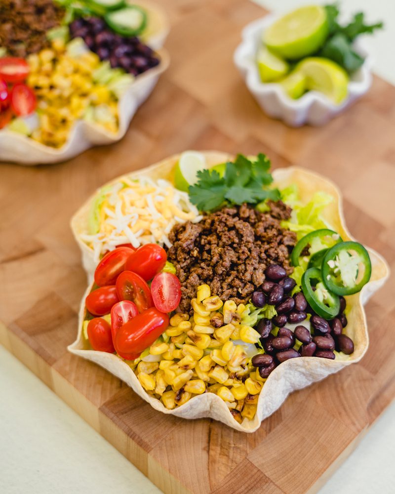 A hearty taco salad served in a crisp tortilla bowl. The salad is topped with seasoned ground beef, black beans, corn, cherry tomatoes, shredded cheese, lettuce and fresh cilantro. Lime wedges are arranged beside the bowl on a wooden cutting board.