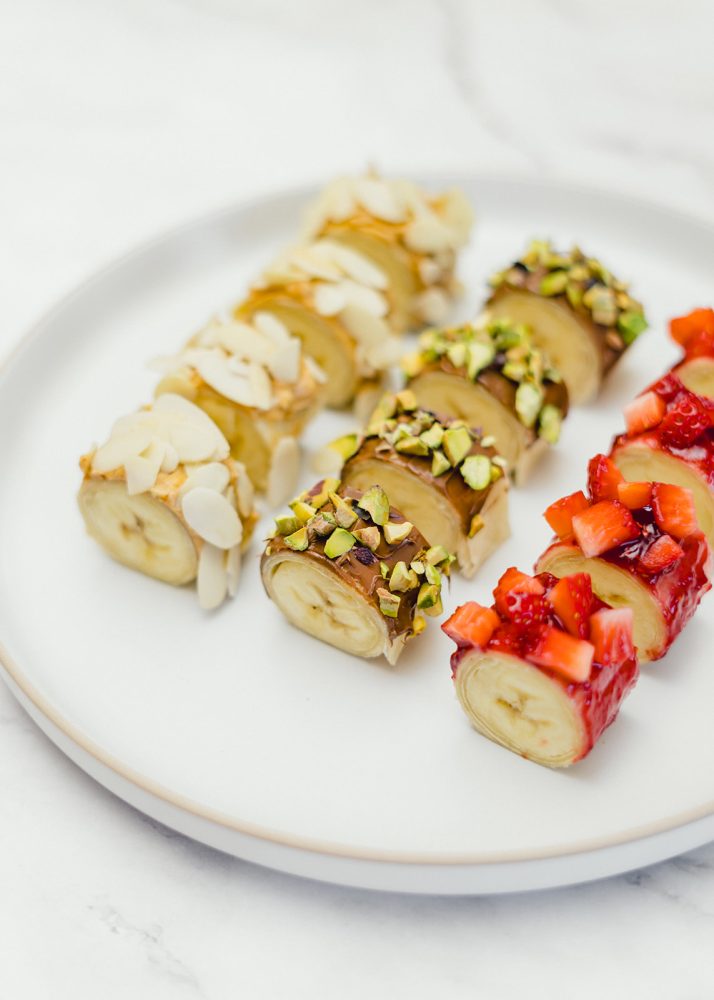 Une assiette blanche contient des sushis sucrés faits de rondelles de banane enrobées d'un pain tortillas. Le pain est ensuite garni de chocolat et de pistaches hachées, de beurre d’arachide et d’amandes effilées ou de confiture aux fraises avec des morceaux de fraises.