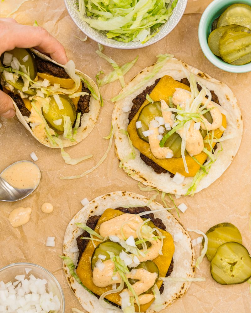 One hand holds a taco filled with a ground beef patty, cheese, pickles, onions and creamy pink/orange sauce. Three similar tacos are arranged on a brown paper surface, with additional ingredients in bowls around them: shredded lettuce, sliced pickles and chopped onions. There's also a spoonful of special sauce.