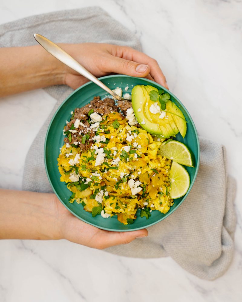 One person holds a green bowl containing scrambled eggs, crushed black beans, feta cheese, avocado and lime slices, garnished with fresh cilantro.