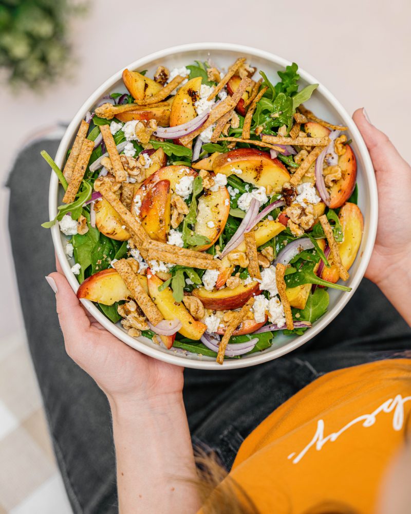 A person wearing an orange T-shirt holds a large white bowl filled with a salad of peaches, arugula, feta cheese, red onions, walnuts and tortilla chips.