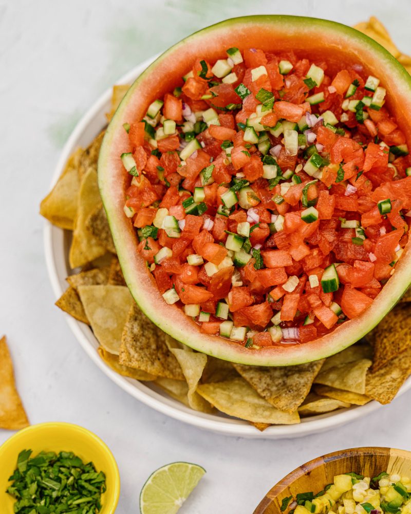 A hollowed-out half-watermelon filled with a refreshing salsa of diced watermelon, tomatoes, cucumbers and red onions. Corn chips surround the watermelon on a white plate. Side dishes include lime wedges, chopped fresh cilantro and a bowl of corn salsa.