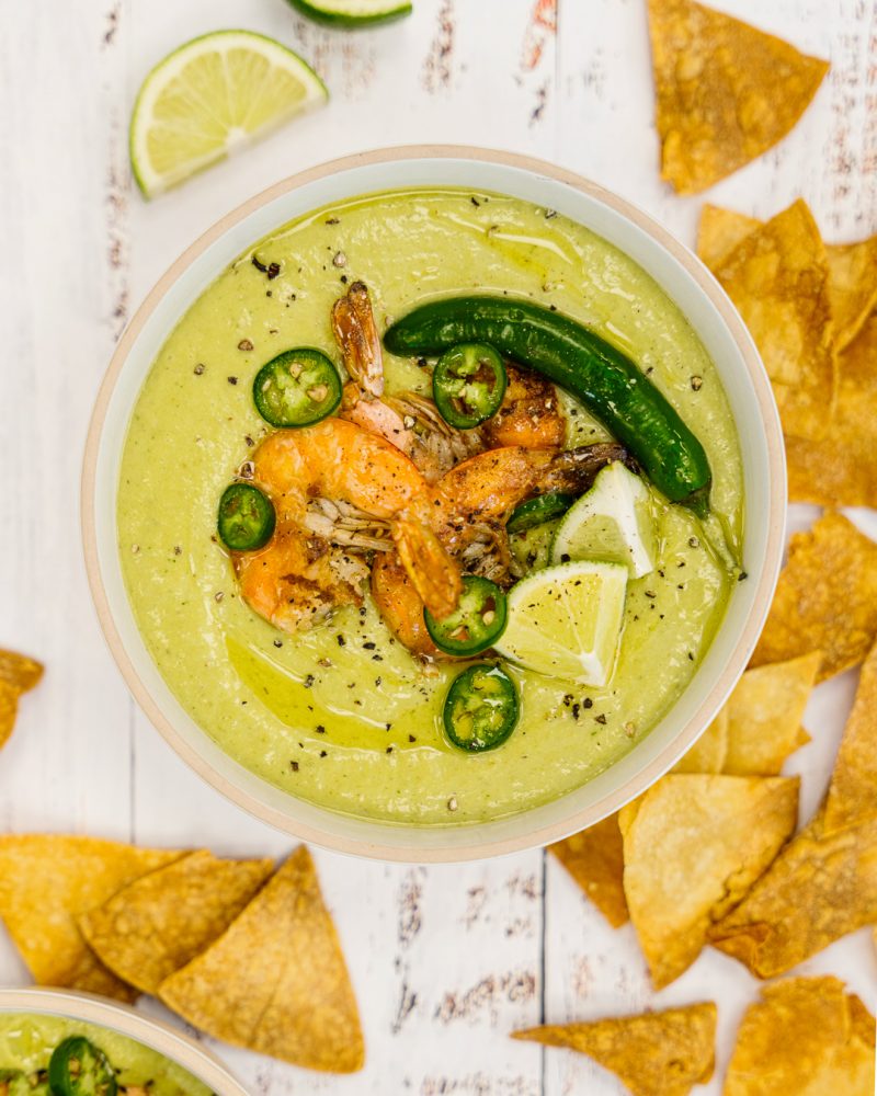 A bowl of creamy guacamole, topped with grilled shrimp, sliced jalapeño and lime, surrounded by tortilla chips.