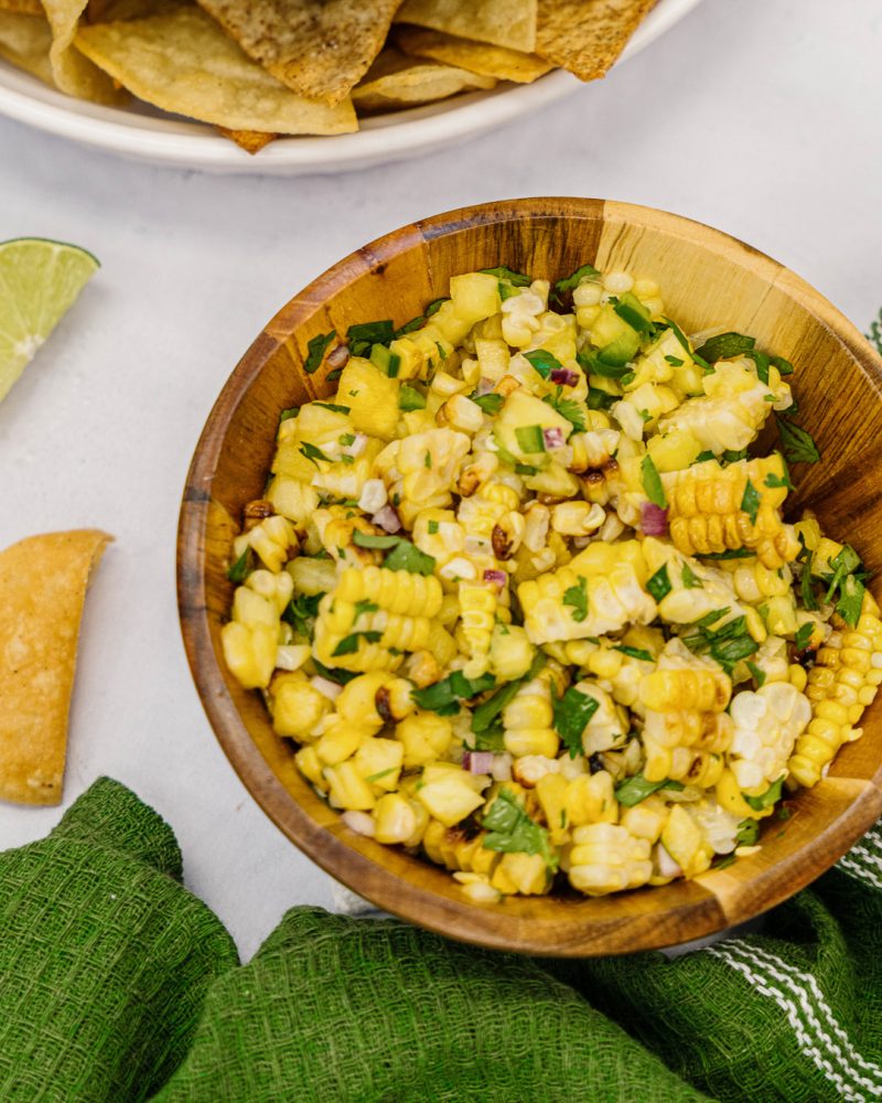 A wooden bowl filled with grilled corn salad with red onions and cilantro. Lime wedges and corn chips are arranged beside the bowl.