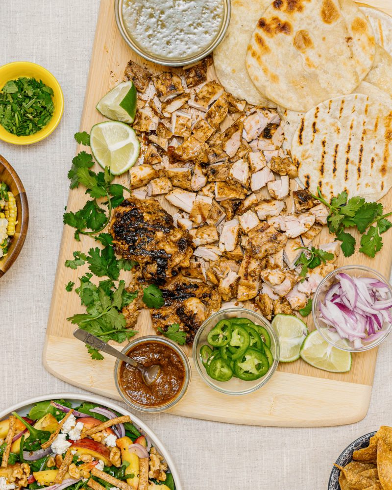 A wooden board filled with grilled chicken and tortillas, surrounded by taco ingredients like salsa, jalapeños, cilantro, red onions, lime wedges and salad.