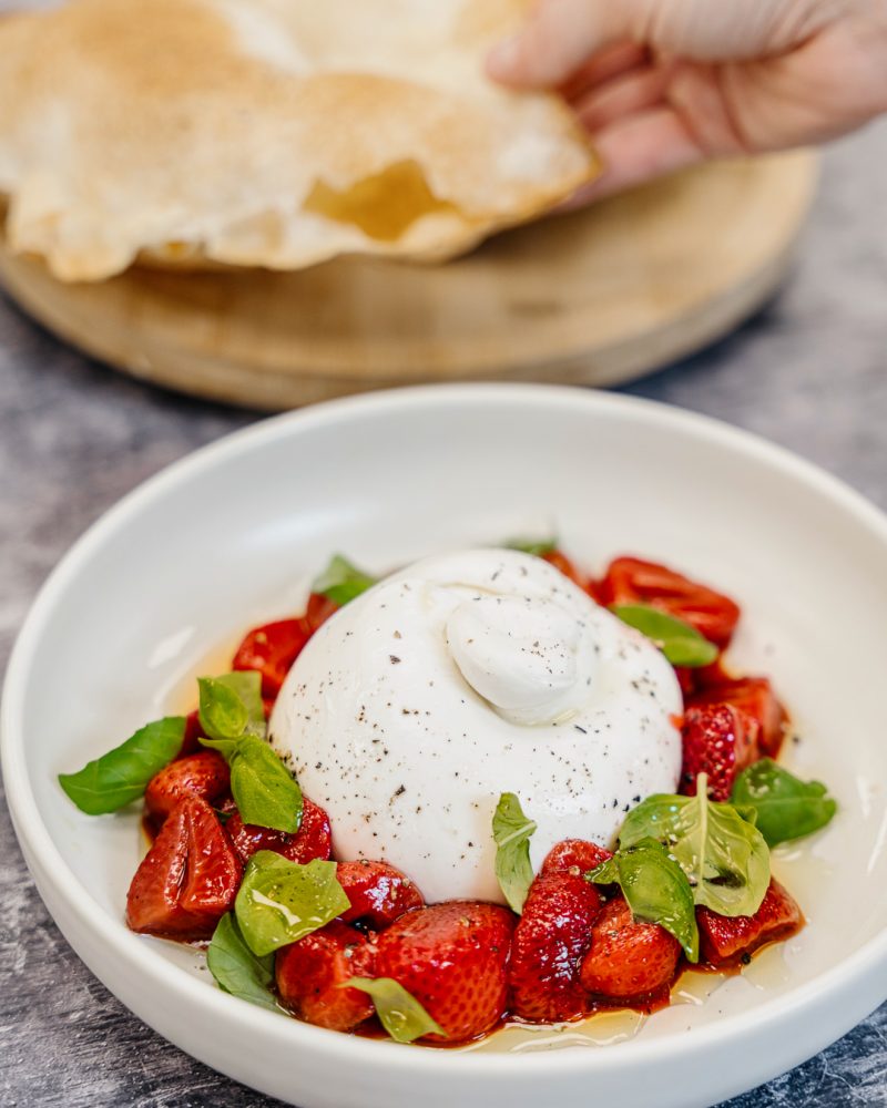 A plate containing creamy burrata, juicy strawberries and fresh basil leaves. In the distance, a hand holds out a crisp tortilla.