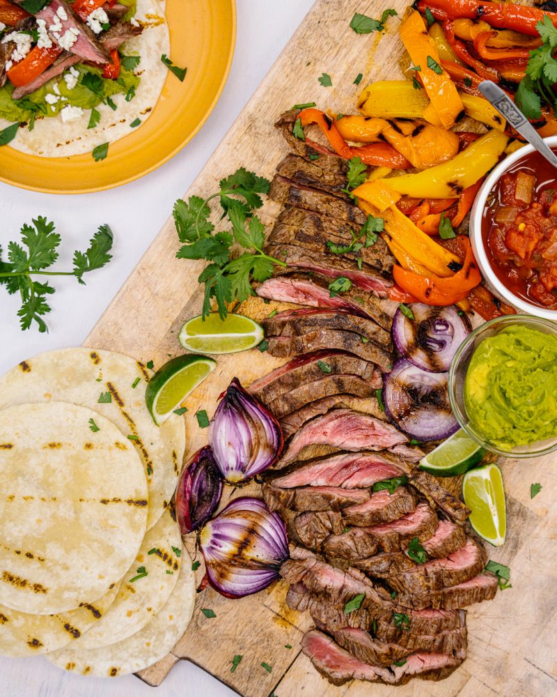 A wooden cutting board with slices of grilled steak, different-colored peppers, grilled red onions, lime wedges and coriander leaves. Two small bowls hold guacamole and salsa. To one side, a yellow plate holds tortillas and a taco already prepared with steak, guacamole and cheese.