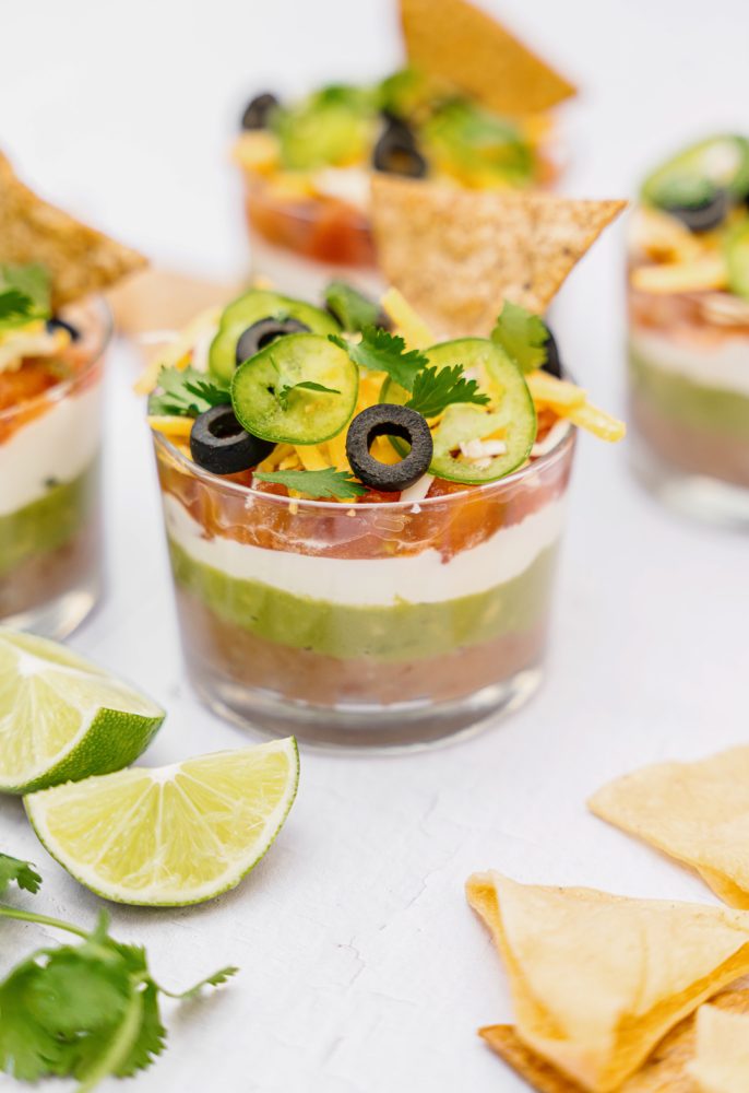 Several small glasses containing a Mexican dip layered with guacamole, sour cream, salsa, shredded cheese, sliced black olives, sliced jalapeño peppers and fresh cilantro. There are lime wedges, tortilla chips and fresh cilantro around the glasses.