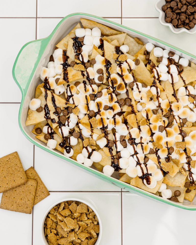 A plate of nachos s'mores in a green baking dish. The nachos are topped with toasted marshmallows, chocolate chips and chocolate sauce. Two small bowls on the side hold graham cracker crumbs and chocolate chips.