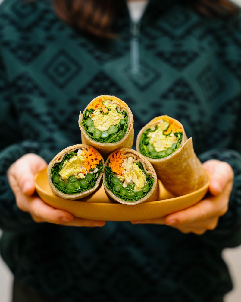 One person holds a plate containing three wraps cut in half and stuffed with eggs salad, asparagus, lettuce and grated carrots.