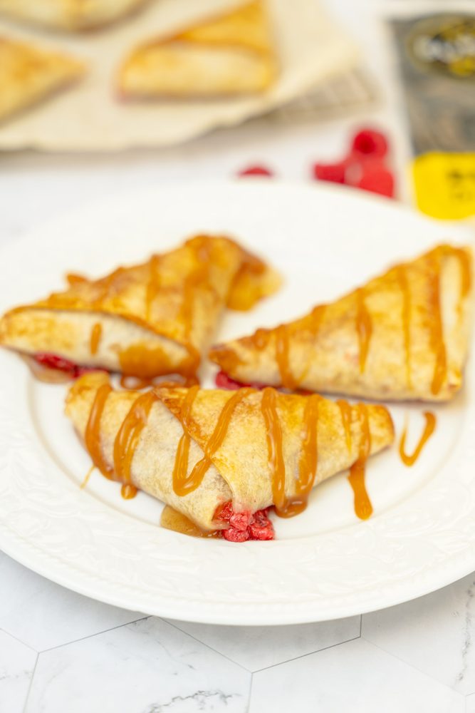 Three triangular pastries filled with raspberries and drizzled with caramel on a white plate. More pastries and raspberries are visible in the background.