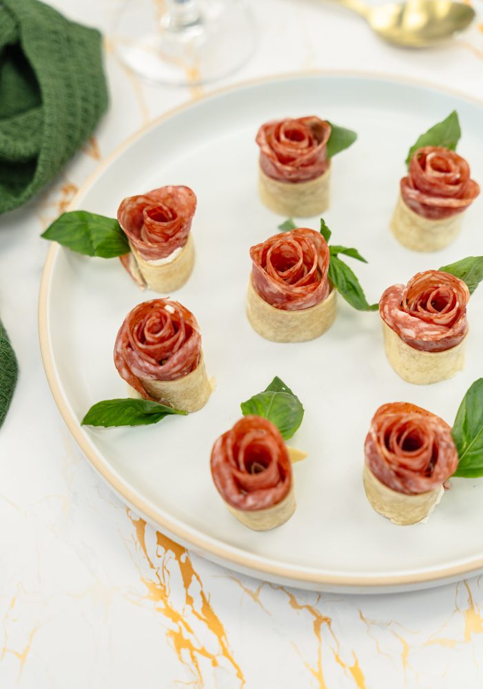 A round white plate displays unique appetizers: thin slices of salami rolled into the shape of roses and placed on small discs of crispy puff pastry. Each salami rose is garnished with a fresh basil leaf for a touch of green color and freshness. The plate is set on a white marble surface with gold accents, and a corner of a green napkin is visible on the left.