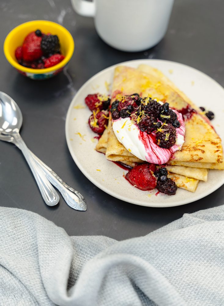 Two thin, golden crêpes are folded into triangles and arranged on a rustic white plate. They are topped with a generous dollop of smooth whipped cream and a medley of fresh, colorful berries, including strawberries, blackberries, and raspberries. In the background, a small yellow bowl filled with more berries, two silver spoons with ornate handles, and a gray linen napkin can be seen. The entire scene is set against a dark background, which accentuates the brightness of the dish.