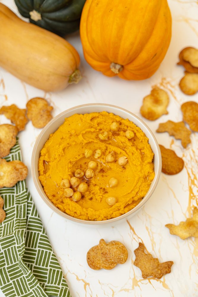 A bowl of creamy, vibrant orange pumpkin hummus, topped with whole chickpeas, a drizzle of olive oil, and a sprinkle of spices. The bowl is surrounded by festive pumpkin and bat-shaped crackers, with a pumpkin, butternut squash, and acorn squash in the background, creating a festive fall scene.