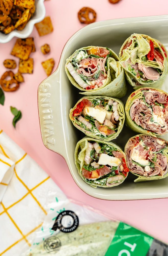 Several wraps cut in half, arranged in a rectangular dish. The wraps contain various ingredients such as prosciutto, mozzarella cheese, vegetables, and a creamy sauce. A package of tortillas, pretzels, and a bowl of chips are also visible on a pink background.