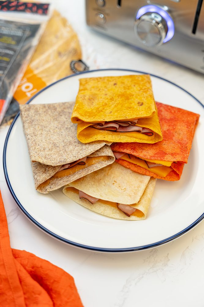 Une assiette blanche avec un bord bleu contient une pile de quesadillas colorées pliées en deux. Les quesadillas sont garnies de jambon et de fromage, et les tortillas sont de différentes couleurs, notamment jaune, rouge, blanc et brun. Un grille-pain et des emballages de tortillas sont visibles en arrière-plan.