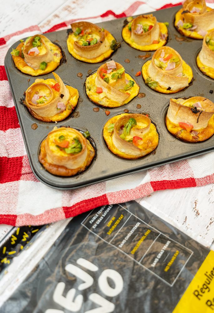 A muffin tin filled with vegetable mini-quiches. The quiches are made with rolled tortillas, filled with eggs and colorful vegetables like red bell peppers, broccoli, and onions. A package of Mejicano tortillas is placed next to it, suggesting it was used in the recipe.