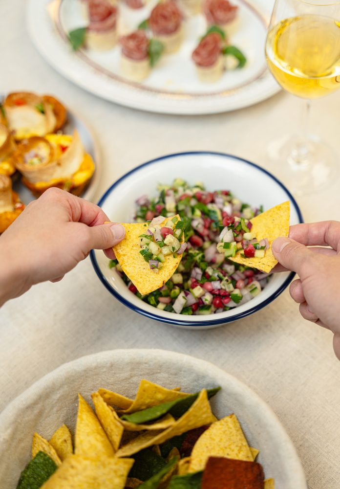 Two hands reach for tortilla chips to scoop up a fresh and colorful pico de gallo from a white bowl. The pico de gallo is a vibrant mix of chopped red onions, green cilantro, juicy red tomatoes, and bright red pomegranate seeds. In the background, other appetizer dishes are visible, including salami roses and mini quiches, along with a glass of white wine. The atmosphere is festive and convivial, suggesting a cocktail party or buffet setting.