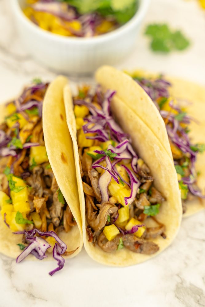 Three tacos filled with shredded mushrooms, pineapple, red cabbage, and cilantro, resting on a marble surface. A bowl of pineapple salsa is visible in the background.