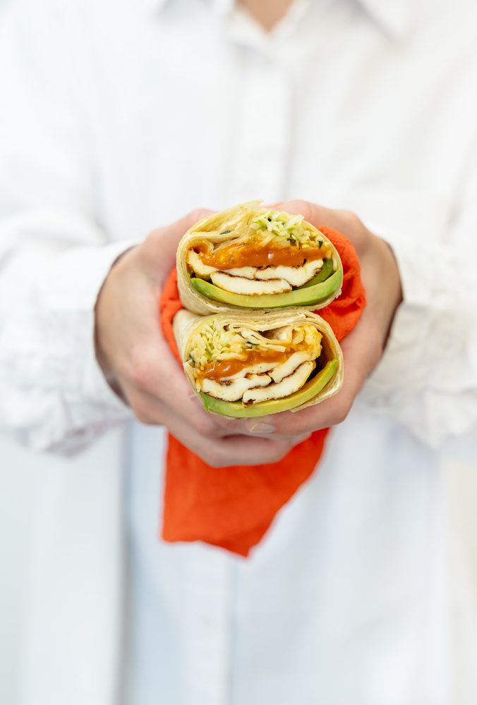 Close-up of two halves of a wrap cut in half, held in an orange napkin. The wrap contains scrambled eggs, cheese, green vegetables, and avocado slices.