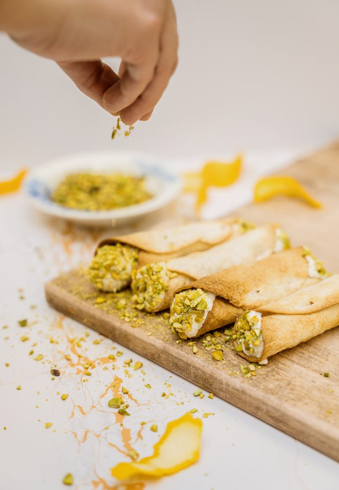 Une main saupoudre des pistaches hachées sur trois cannoli disposés sur une planche à découper en bois. Les cannoli sont garnis d'une crème pâle et de pistaches. Des zestes d'orange sont visibles sur la surface autour de la planche.