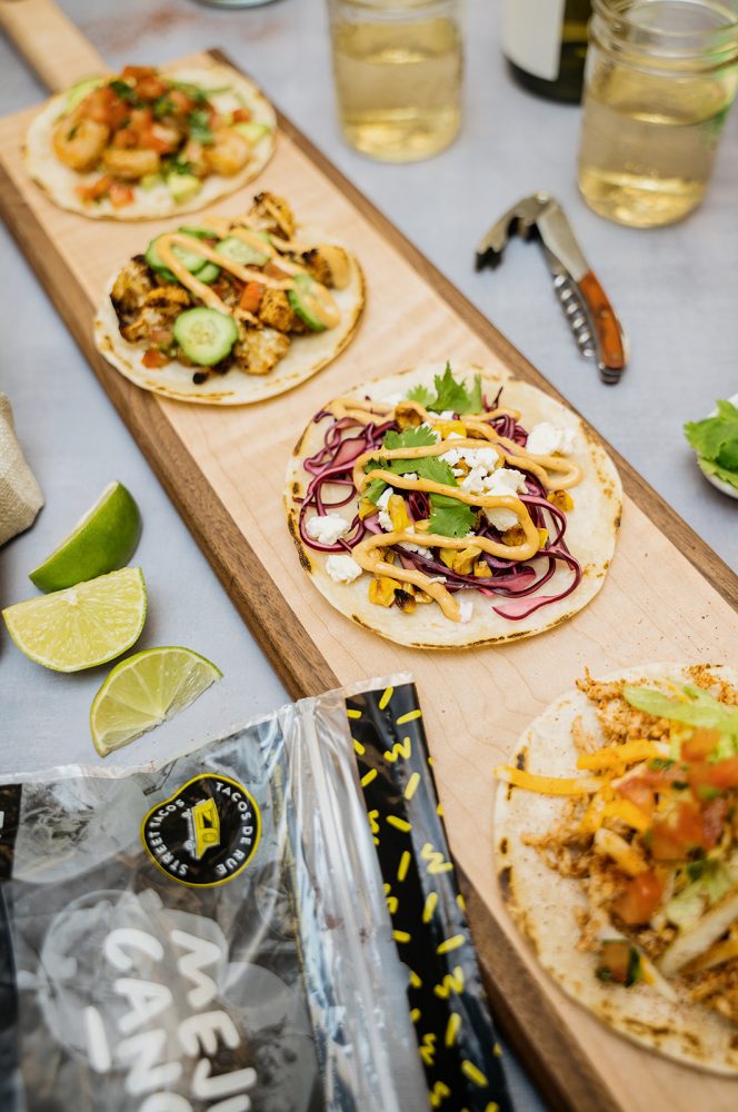 Quatre tacos différents présentés sur une planche de bois, garnis de garnitures colorées comme de la salsa, du fromage et de la coriandre. Des quartiers de lime et un sac de tortillas Mejicano accompagnent les tacos.