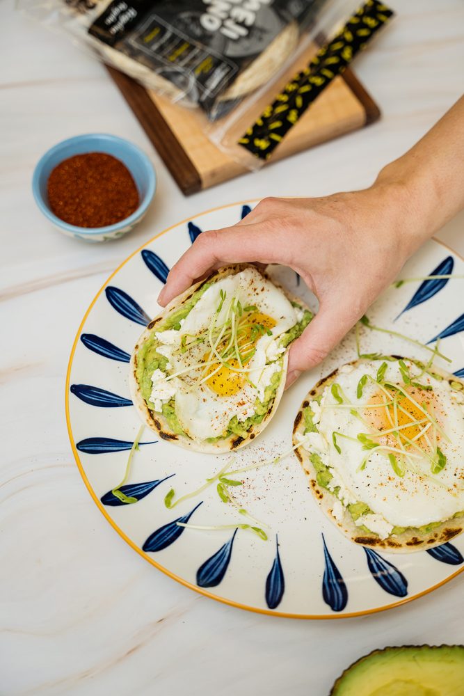 Hand holding a breakfast taco with egg, feta, avocado, and sprouts, served on a blue plate with Mejicano tortillas and a bowl of chili powder.