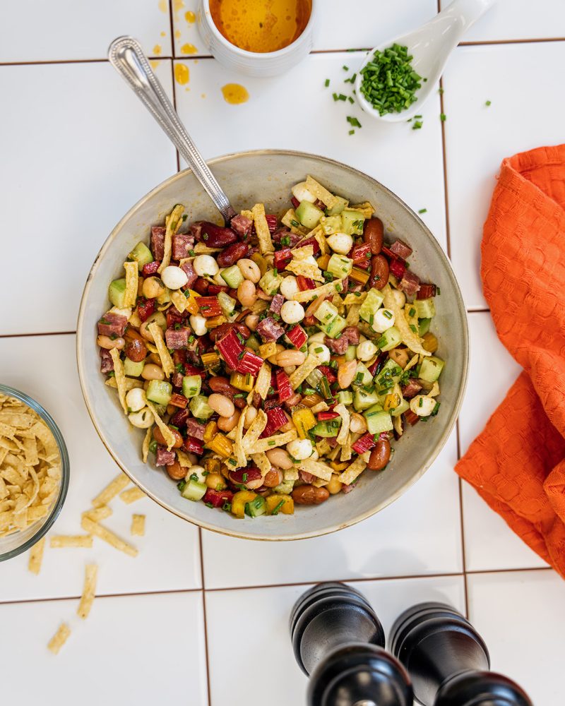 Dense bean salad with red beans, legumes, cucumber, celery, and charcuterie, topped with crispy Mejicano tortilla pieces, served in a bowl with a spoon, orange napkin, chives, salt shaker, and pepper grinder.