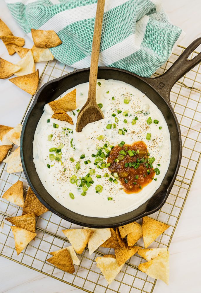 A cast iron skillet filled with creamy white queso dip, topped with chopped green onions and a dollop of salsa. Crispy tortilla triangles are arranged around the skillet on a gold cooling rack.