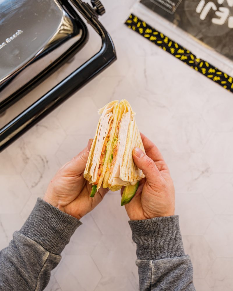 Main tenant un wrap panini inspiré de TikTok avec dinde, tomate et avocat, servi avec tortillas Mejicano et une presse à panini en arrière-plan.