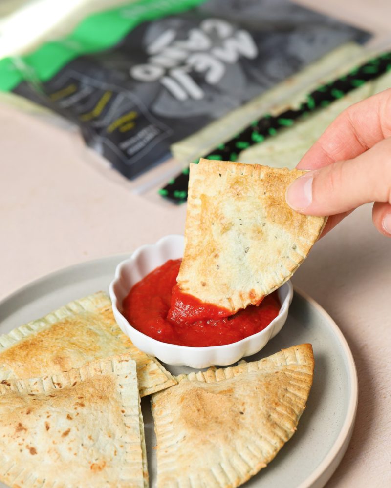 mejicano_tortillas_pesto_raviolis_tofu_epinards_sauce_tomate Trois empanadas triangulaires et dorées sur une assiette grise, accompagnées d’un petit bol blanc rempli de sauce tomate rouge. Une main trempe un morceau d’empanada dans la sauce. Un sac de tortillas est visible en arrière-plan.