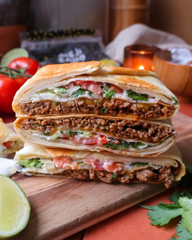 Un wrap croustillant coupé en deux et empilé, révélant des couches de bœuf haché, de fromage fondu, de tomates, de laitue et de crème sure entre deux tortillas croustillantes et une grande tortilla souple. Il repose sur une planche de bois avec de la coriandre fraîche et des quartiers de lime.