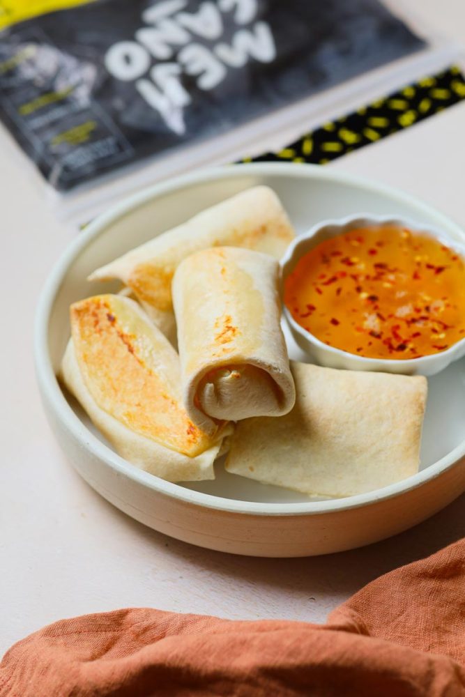 mejicano_tortillas_rouleaux_feta_miel_epice-800X1200 Une assiette blanche contient quatre rouleaux de tortillas grillés, avec une petite coupelle de sauce au miel orange parsemée de flocons de piment. Un emballage noir avec du texte partiellement visible est en arrière-plan.