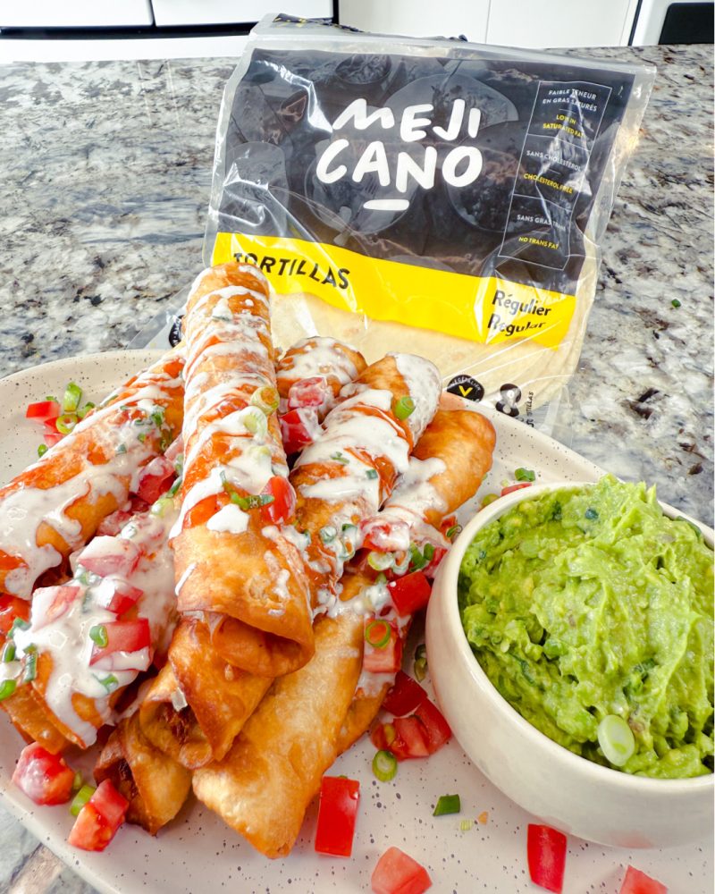 A plate of fried taquitos, drizzled with cream, diced tomatoes, and chopped green onions, served with a bowl of guacamole. A package of Mejicano tortillas is visible in the background.