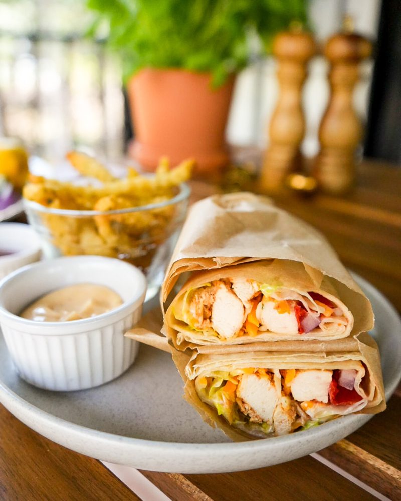 Deux wraps coupés en deux, révélant une garniture de poulet grillé, de laitue, de fromage râpé et de tomates tranchées, sont présentés sur une assiette blanche avec un bol de frites et une trempette. L'arrière-plan flou montre une plante verte et des salières et poivrières en bois.