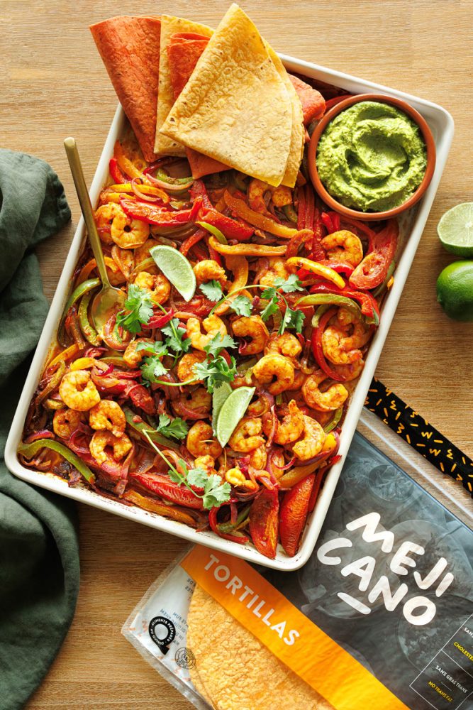 Une vue de dessus d'un grand plat de fajitas aux crevettes et légumes. Le plat rectangulaire blanc est rempli de crevettes roses cuites, de lanières de poivrons rouges et verts, d'oignons et de tomates, le tout assaisonné d'épices. Il est garni de quartiers de lime et de feuilles de coriandre fraîche. Dans le coin supérieur droit du plat, il y a un petit monticule de tortillas coupées en triangles. À côté du plat, se trouvent un bol de guacamole vert et une paire de limes entières. Un paquet de tortillas et un paquet d'épices, tous deux de la marque "MEJICANO", sont également visibles sur une table en bois.