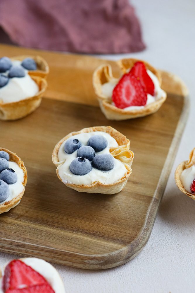 Plusieurs mini-tartelettes sont présentées sur un plateau en bois. Elles sont faites de croûtes croustillantes et remplies d'une crème blanche au fromage. Certaines sont garnies de bleuets entiers, tandis que d'autres sont garnies de fraises tranchées. Un tissu violet est légèrement visible en arrière-plan.