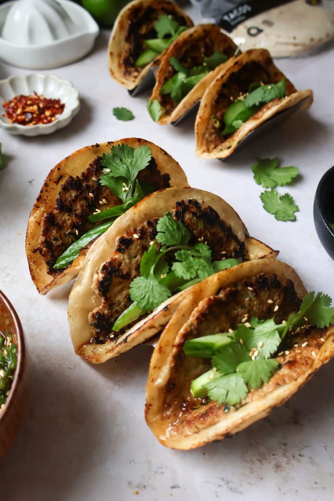 Une série de six tacos cuits à la poêle avec des coquilles dorées et croustillantes sont garnis de viande hachée, de feuilles de coriandre fraîche et de tranches de concombre ou d'avocat. Ils sont disposés sur une surface claire avec divers petits bols d'accompagnement flous en arrière-plan.