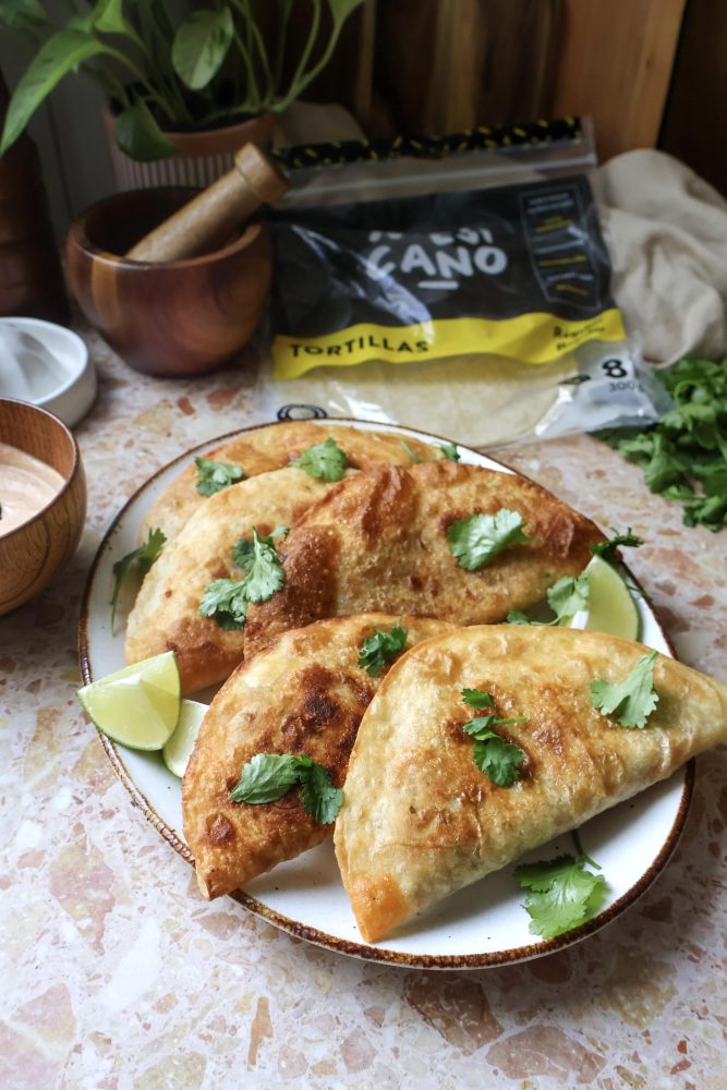 Une assiette de trois empanadas dorés et croustillants, garnis de feuilles de coriandre fraîche et de quartiers de lime, est présentée sur un comptoir en marbre. En arrière-plan, on aperçoit un bol de sauce, un pilon et un mortier, une plante en pot et un paquet de tortillas Mejicano.