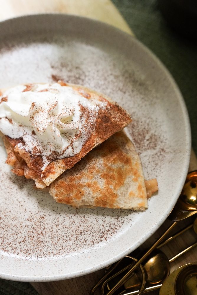 mejicano_tortillas_wraps_pommes_miel_fromage_fondant Une crêpe ou tortilla pliée, garnie d'une généreuse portion de crème fouettée et saupoudrée de cannelle ou de cacao en poudre, est posée sur une assiette beige. La poudre est également répandue autour de la crêpe sur l'assiette. Des cuillères à mesurer dorées sont partiellement visibles dans le coin inférieur droit.