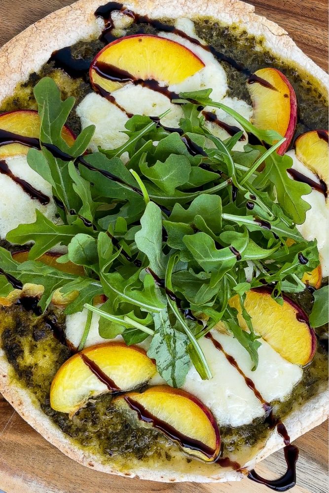An appetizing pizza is displayed on a crispy crust, spread with a vibrant green pesto base. It's topped with slices of creamy burrata cheese, juicy peach wedges, and a generous pile of fresh arugula. A drizzle of dark balsamic glaze completes the dish.