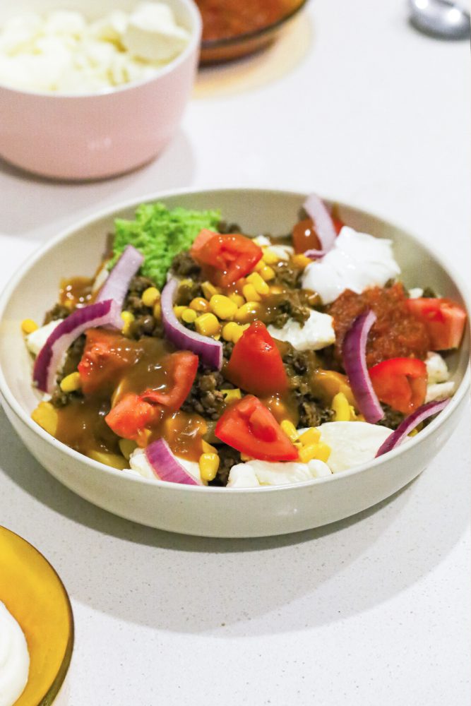 A creative poutine is served in a bowl, featuring a base of french fries topped with cheese curds, ground meat, corn, diced tomatoes, sliced red onions, and brown gravy. It's garnished with a dollop of sour cream and guacamole. In the blurred background, a pink bowl of cheese curds and a spoon are visible.