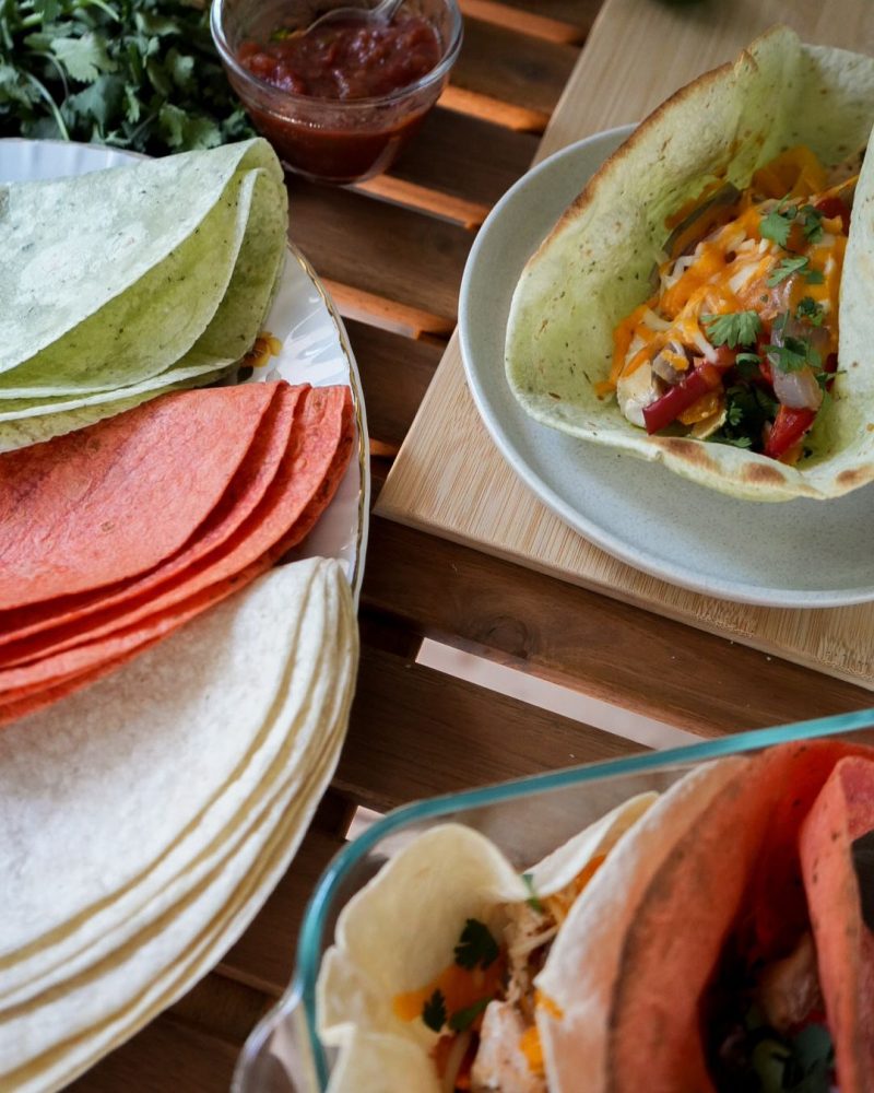 Une table en bois garnie de tortillas Mejicano colorées (rouges, vertes et blanches), de salsa, coriandre fraîche, lime, et de tortillas farcies de légumes grillés, poulet et fromage râpé. Une ambiance conviviale et festive de repas à partager.